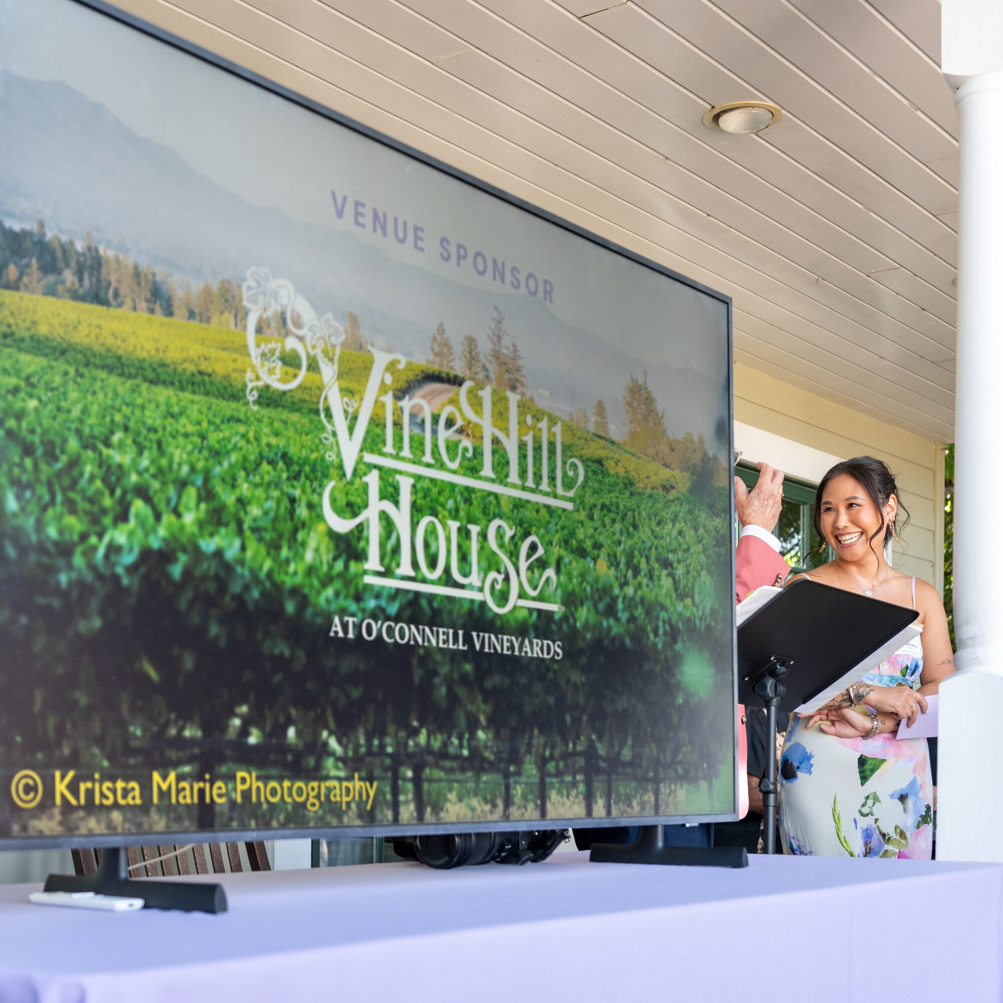A TV displays the Vine Hill House logo while a women beside it smiles widely