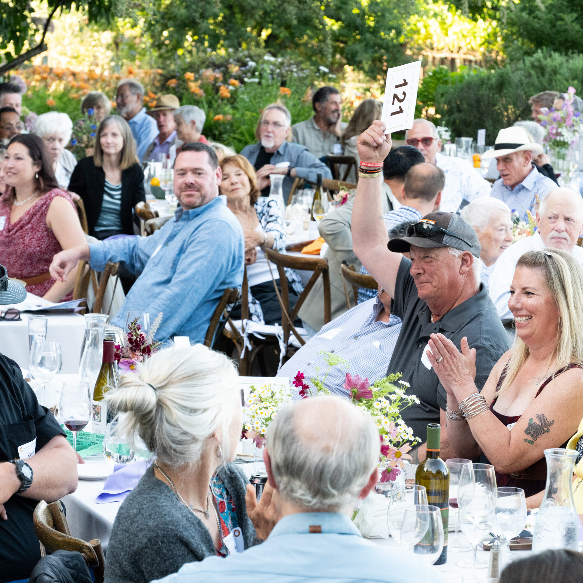 A man raises his auction bidder number while guests around him laugh, clap, and smile